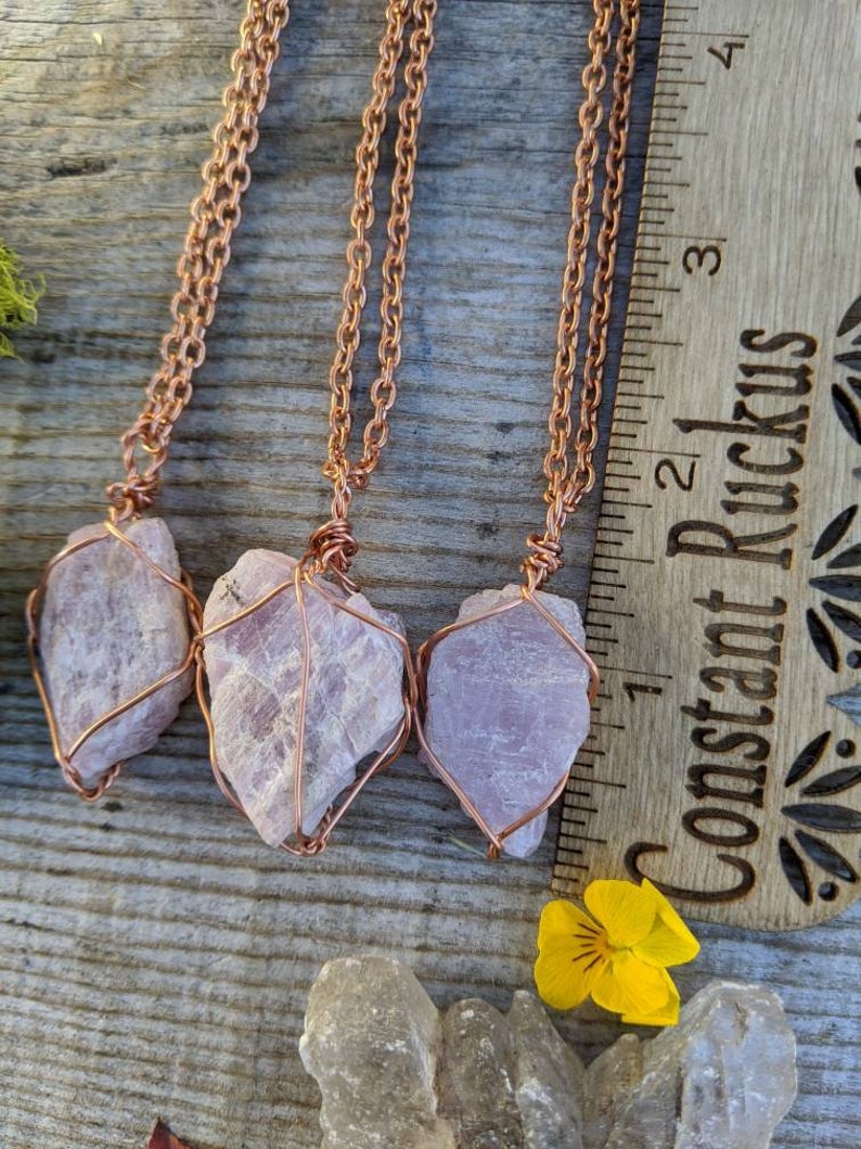 May include: Three pink crystal pendants wrapped in copper wire, hanging from copper chains. The pendants are approximately 1 inch long and are displayed on a wooden surface with a ruler.