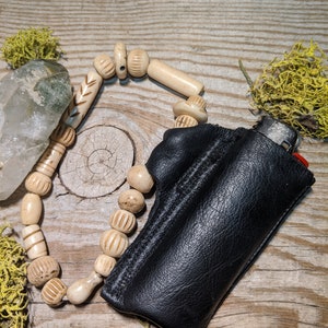 Puede incluir: Una funda de cuero negro para encendedor con un collar de cuentas de hueso blanco. La funda tiene una solapa que se cierra con un broche a presi&oacute;n. El collar est&aacute; hecho de cuentas redondas con intrincados tallados.