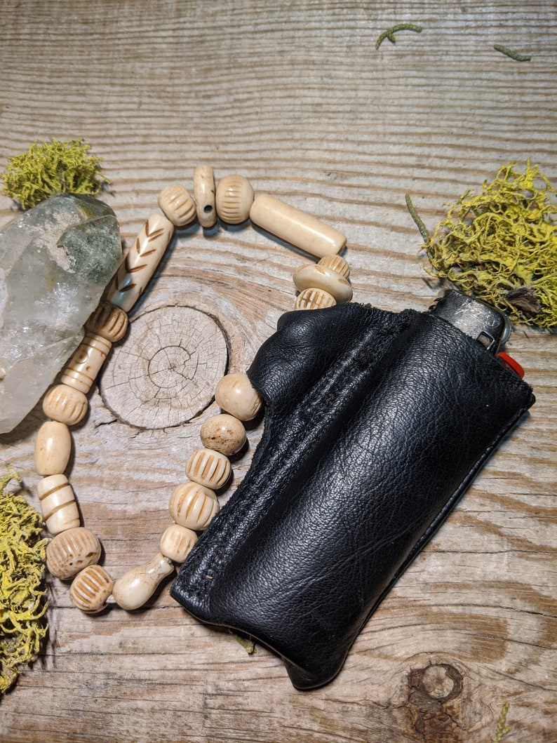 Puede incluir: Una funda de cuero negro para encendedor con un collar de cuentas de hueso blanco. La funda tiene una solapa que se cierra con un broche a presi&oacute;n. El collar est&aacute; hecho de cuentas redondas con intrincados tallados.
