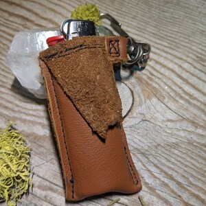 May include: A brown leather keychain with a small pocket for a lighter. The keychain has a silver metal loop and a black stitching detail.