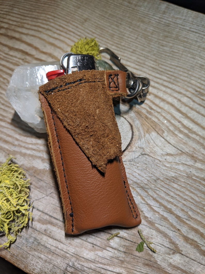 May include: A brown leather keychain with a small pocket for a lighter. The keychain has a silver metal loop and a black stitching detail.