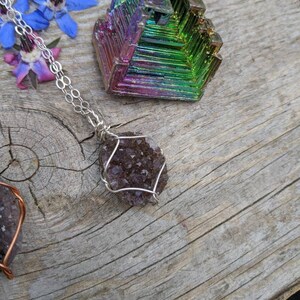 May include: A silver chain necklace with a purple amethyst crystal pendant wrapped in silver wire. The crystal is a cluster of small, faceted stones. The necklace is displayed on a wooden surface with a colorful iridescent crystal.