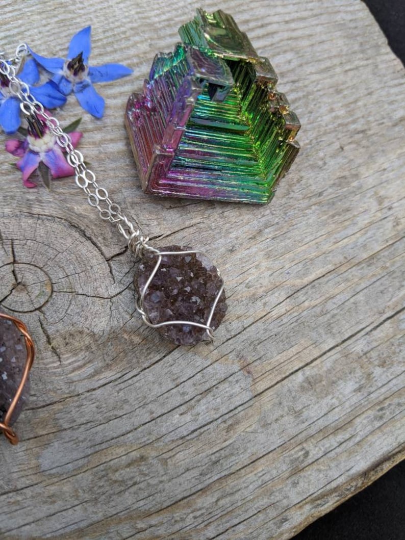May include: A silver chain necklace with a purple amethyst crystal pendant wrapped in silver wire. The crystal is a cluster of small, faceted stones. The necklace is displayed on a wooden surface with a colorful iridescent crystal.