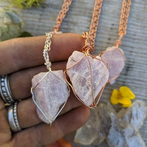May include: Three necklaces with pink crystal pendants wrapped in wire. The necklaces are on a wooden surface with moss and a yellow flower.