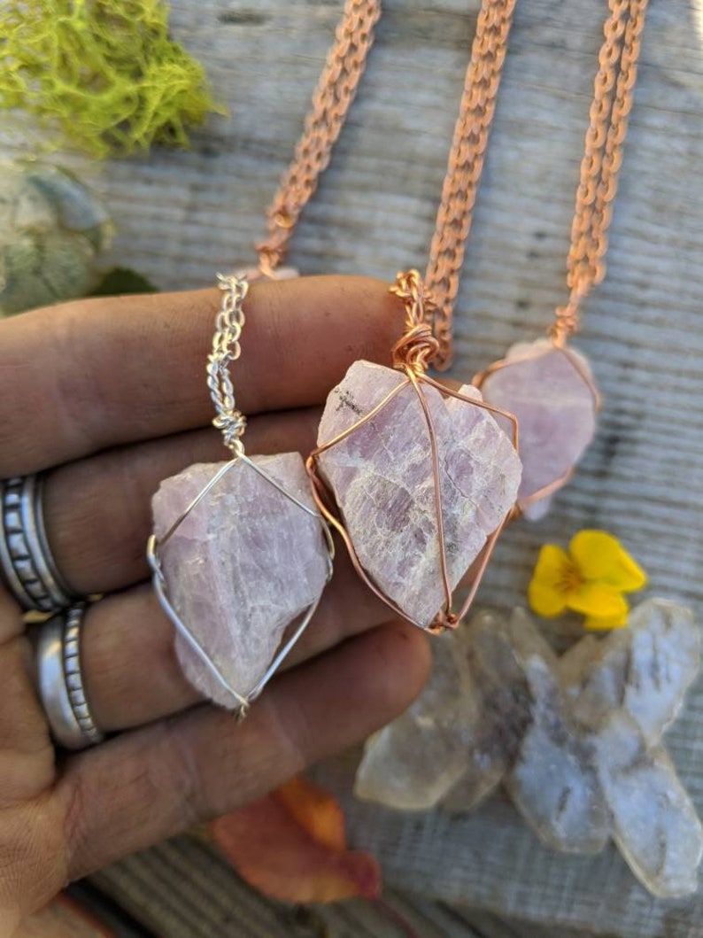 May include: Three necklaces with pink crystal pendants wrapped in wire. The necklaces are on a wooden surface with moss and a yellow flower.