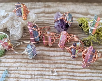Puede incluir: Una colección de anillos envueltos en alambre con puntas de cristal iridiscentes en varios colores, incluyendo rosa, morado y azul. Los anillos están hechos con alambre de cobre y se muestran sobre una superficie de madera.