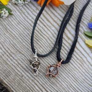 Puede incluir: Dos collares con colgantes de cristal marr&oacute;n envueltos en alambre. Un colgante est&aacute; envuelto en alambre plateado y el otro en alambre de cobre. Los collares est&aacute;n sobre una superficie de madera con flores en el fondo.