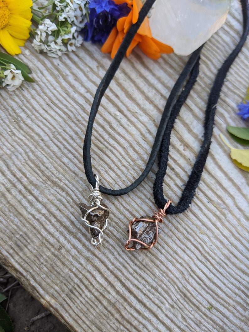 Puede incluir: Dos collares con colgantes de cristal marr&oacute;n envueltos en alambre. Un colgante est&aacute; envuelto en alambre plateado y el otro en alambre de cobre. Los collares est&aacute;n sobre una superficie de madera con flores en el fondo.