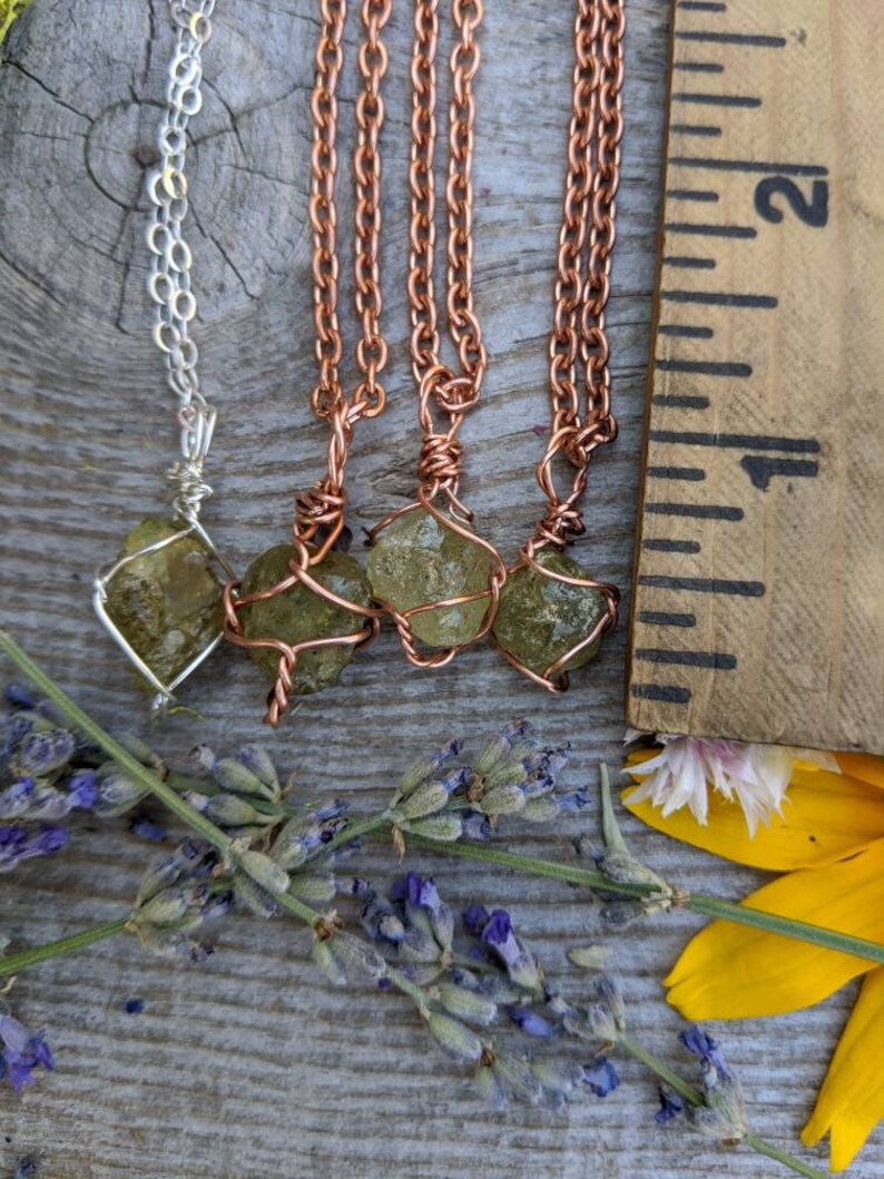 May include: Four necklaces with green gemstone pendants wrapped in copper wire. The necklaces are on a wooden surface with lavender flowers and a yellow flower.