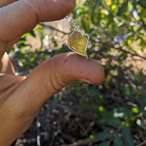 May include: A yellow crystal pendant wrapped in silver wire. The pendant is being held by a hand.