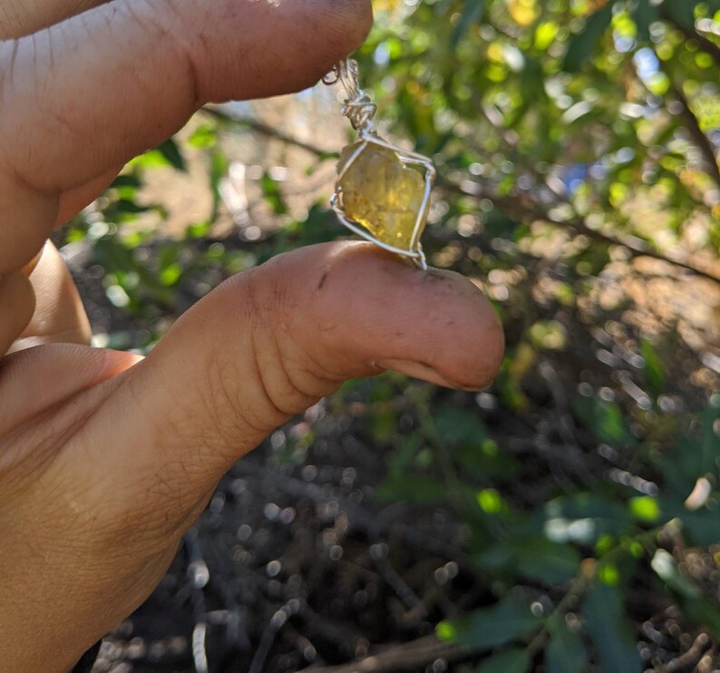 May include: A yellow crystal pendant wrapped in silver wire. The pendant is being held by a hand.
