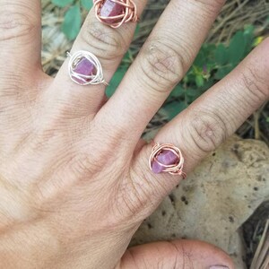 May include: Two wire wrapped rings with pink gemstones. The rings are made with copper wire and feature a pink gemstone in the center. The rings are shown on a hand.
