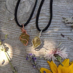 May include: Two necklaces with green gemstone pendants wrapped in wire. One pendant is wrapped in copper wire and the other is wrapped in silver wire. The necklaces are on a wooden surface with flowers.