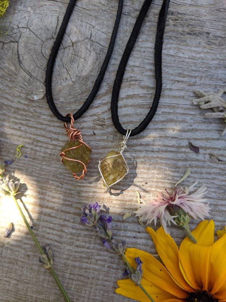 May include: Two necklaces with green gemstone pendants wrapped in wire. One pendant is wrapped in copper wire and the other is wrapped in silver wire. The necklaces are on a wooden surface with flowers.