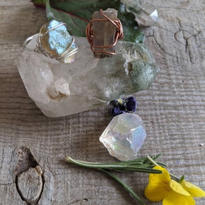 May include: Two wire-wrapped rings with iridescent stones, one with a blue-green stone and one with a brown stone. The rings are displayed on a clear quartz crystal with a green moss agate crystal and a clear quartz crystal. A sprig of rosemary and yellow flowers are in the foreground.
