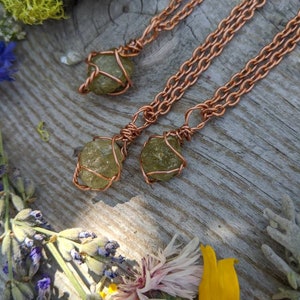 May include: Three copper wire wrapped green gemstone pendants on copper chain necklaces. The necklaces are displayed on a wooden surface with green moss, lavender, and yellow flowers.