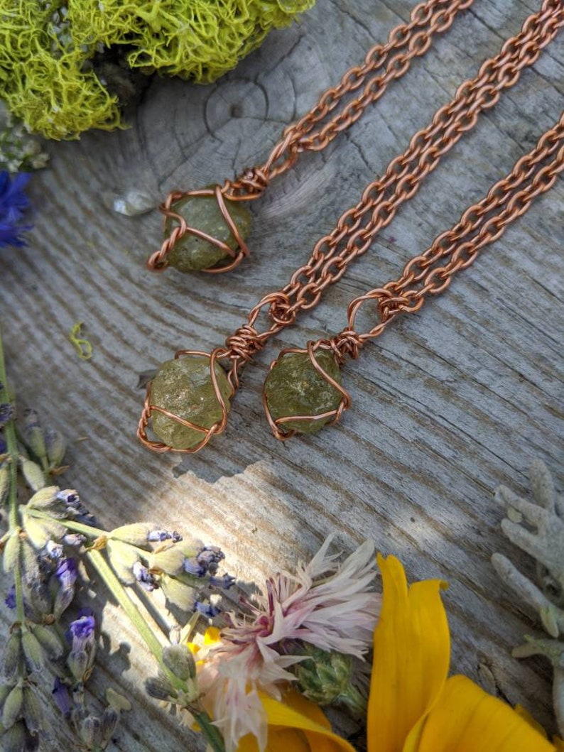 May include: Three copper wire wrapped green gemstone pendants on copper chain necklaces. The necklaces are displayed on a wooden surface with green moss, lavender, and yellow flowers.