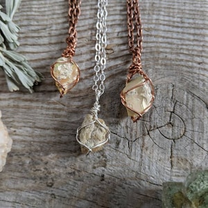 May include: Three necklaces with wire-wrapped, raw, yellow-toned crystal pendants. The necklaces are on a wooden surface. The pendants are suspended from silver and copper chains.