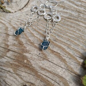 May include: A pair of silver earrings with a blue gemstone dangling from each. The earrings feature a chain with multiple silver rings.