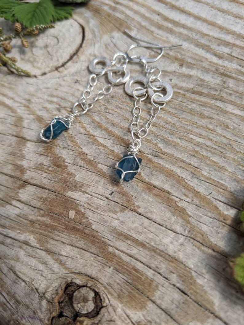 May include: A pair of silver earrings with a blue gemstone dangling from each. The earrings feature a chain with multiple silver rings.