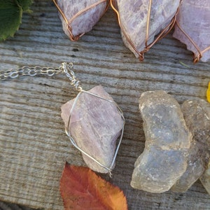 May include: A silver chain necklace with a pink crystal pendant encased in a silver wire cage. The crystal is a natural, rough-cut shape. The necklace is displayed on a wooden surface with other pink crystals wrapped in copper wire.