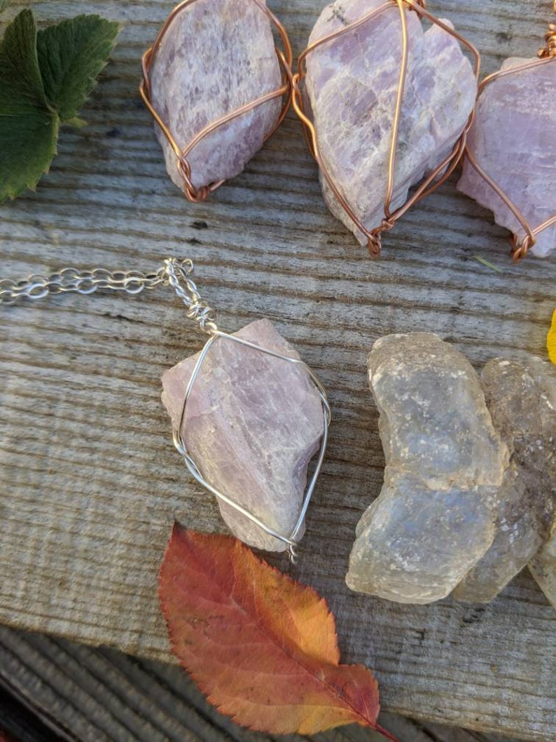 May include: A silver chain necklace with a pink crystal pendant encased in a silver wire cage. The crystal is a natural, rough-cut shape. The necklace is displayed on a wooden surface with other pink crystals wrapped in copper wire.