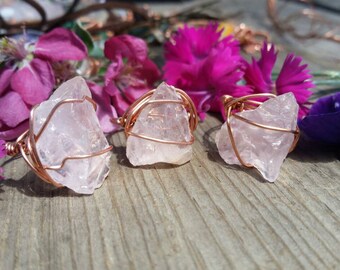 May include: Three rose quartz rings wrapped in copper wire. The rings are displayed on a wooden surface with pink flowers.