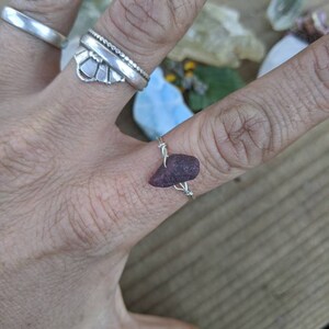 May include: A hand wearing three silver rings, including a wire-wrapped ring with a dark purple gemstone. The other rings are a simple band and a decorative ring with a fan-like design. The background includes other gemstones.