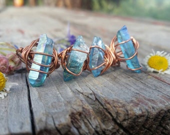 Puede incluir: Cuatro anillos hechos a mano con piedras de cristal azul envueltas en alambre de cobre. Los anillos están colocados sobre una superficie de madera envejecida, con pequeñas flores y una margarita en el fondo. Los cristales son de color azul claro.