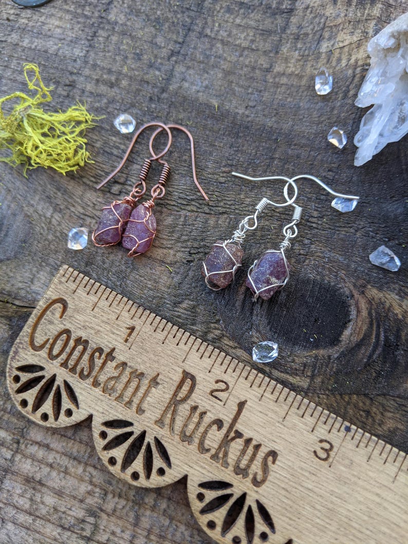 K&ouml;nnte beinhalten: Ein Paar Ohrringe mit rosa und violetten Kristallsteinen, die in Kupfer- und Silberdraht gewickelt sind. Die Ohrringe h&auml;ngen an einem Holzlineal mit der Gravur "Constant Ruckus".