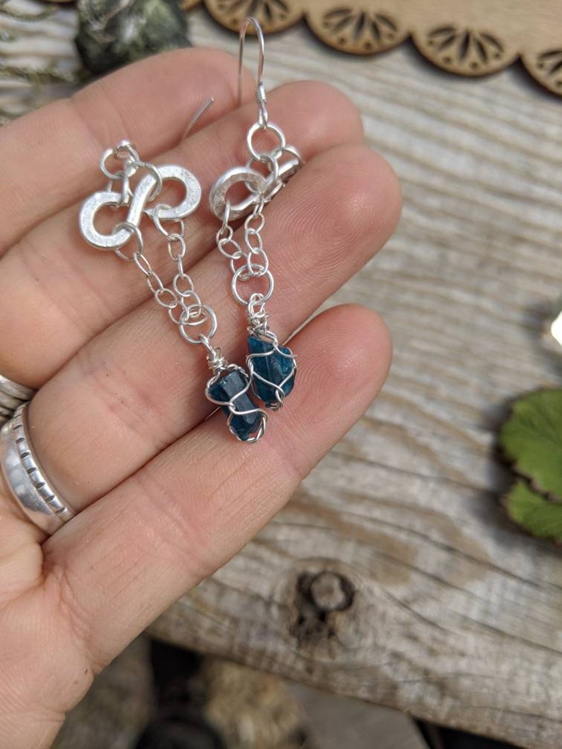 May include: Silver infinity symbol earrings with a dangling chain and a teal blue gemstone at the end.