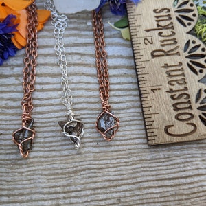 Puede incluir: Tres collares con colgantes de piedra gris oscuro envueltos en alambre. Los colgantes miden aproximadamente 2,5 cm de largo. Los collares est&aacute;n sobre una superficie de madera con una regla etiquetada como "Constant Ruckus" en el fondo.