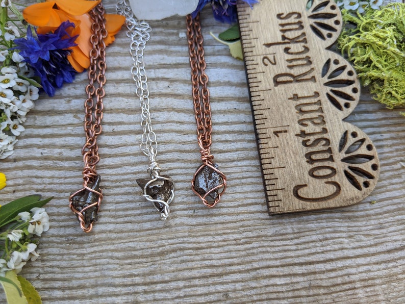 Puede incluir: Tres collares con colgantes de piedra gris oscuro envueltos en alambre. Los colgantes miden aproximadamente 2,5 cm de largo. Los collares est&aacute;n sobre una superficie de madera con una regla etiquetada como "Constant Ruckus" en el fondo.