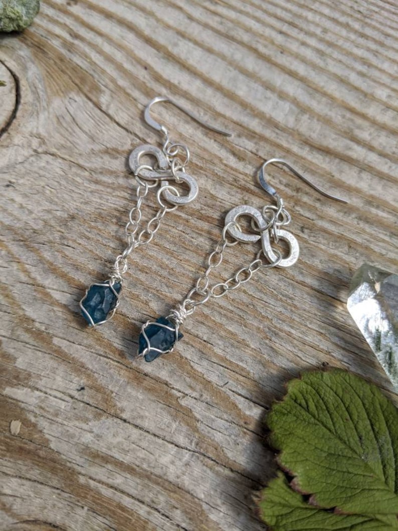 May include: A pair of silver earrings with infinity symbol charms and dangling blue crystal stones.