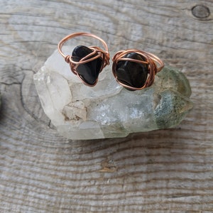 May include: Two handmade rings with black stones, set on a clear quartz crystal. The rings feature copper wire wrapping around the stones and forming the ring band. The background is a weathered wooden surface.