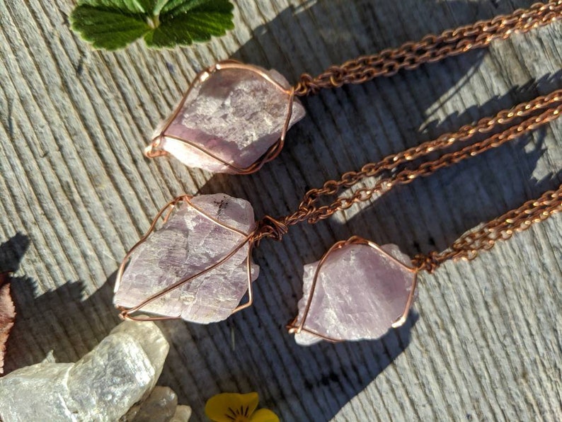 May include: Three pink crystal pendants wrapped in copper wire, hanging from copper chains. The crystals are raw and have a natural, irregular shape.