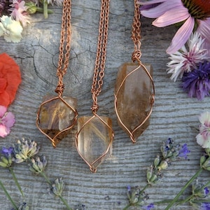 May include: Three gold and brown crystal necklaces with copper wire wrapped around the crystals and copper chains. The necklaces are displayed on a wooden surface with flowers.