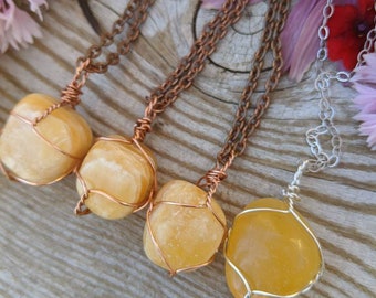 May include: Four necklaces with yellow-orange gemstones. Three have copper chains and wire wrapping, while the fourth features a silver chain and wire. The gemstones are smooth and rounded, with a natural, organic appearance. The background includes flowers.
