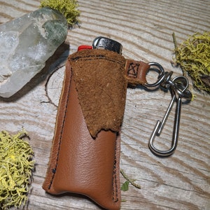May include: A brown leather lighter case with a suede flap and a silver metal keychain. The case is sitting on a wooden surface.