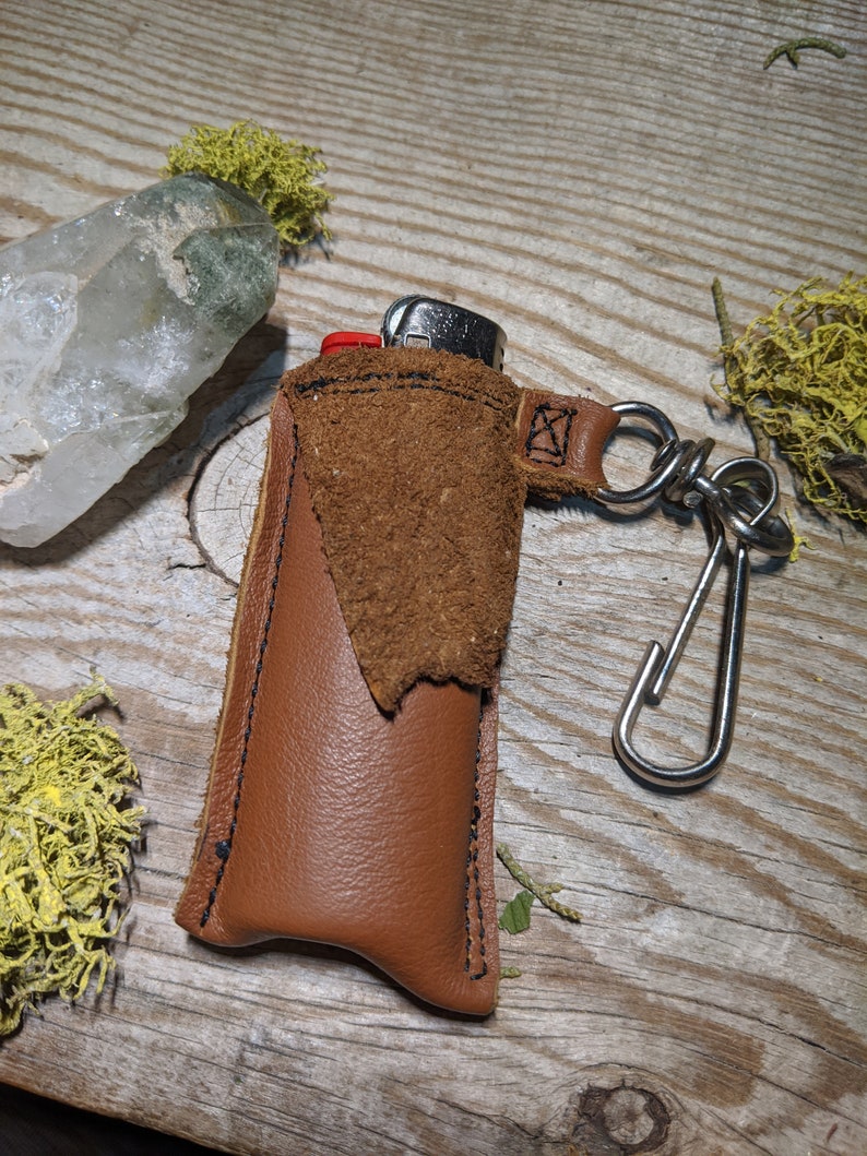 May include: A brown leather lighter case with a suede flap and a silver metal keychain. The case is sitting on a wooden surface.