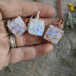 Puede incluir: Tres colgantes de piedra lunar iridiscente envueltos en alambre de cobre con un delicado dise&ntilde;o de coraz&oacute;n. Los colgantes cuelgan de cadenas de cobre.