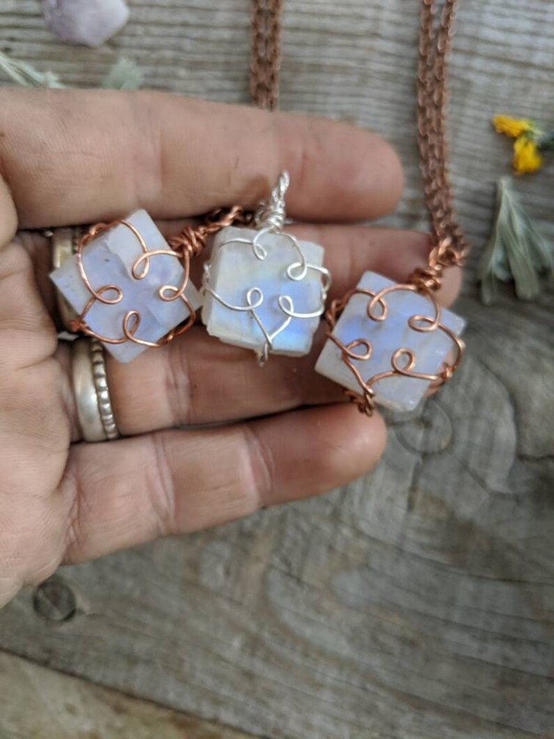 Puede incluir: Tres colgantes de piedra lunar iridiscente envueltos en alambre de cobre con un delicado dise&ntilde;o de coraz&oacute;n. Los colgantes cuelgan de cadenas de cobre.