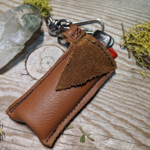 May include: A brown leather lighter case with a suede flap and a metal keychain. The case is stitched with black thread and has a red lighter inside.