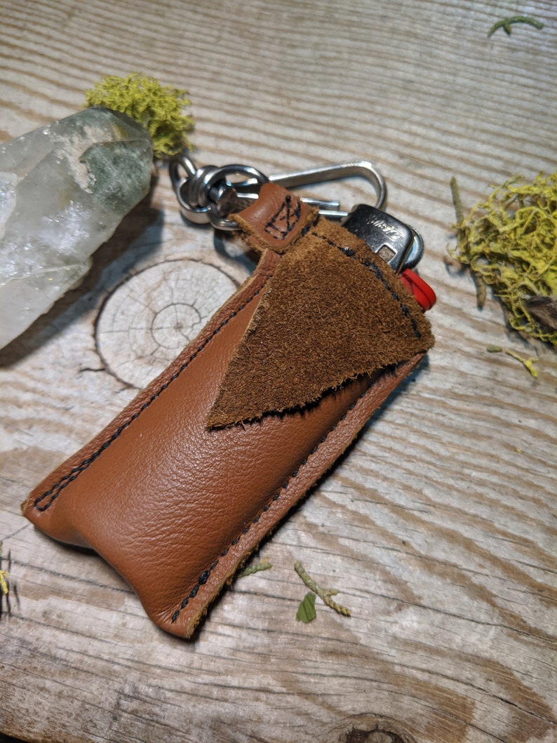 May include: A brown leather lighter case with a suede flap and a metal keychain. The case is stitched with black thread and has a red lighter inside.
