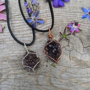 May include: Two amethyst crystal necklaces with copper wire wrapping. The necklaces are on a wooden surface with purple flowers.