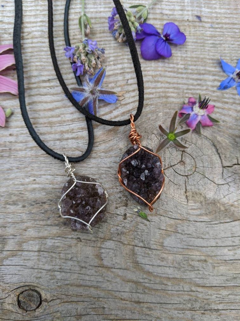 May include: Two amethyst crystal necklaces with copper wire wrapping. The necklaces are on a wooden surface with purple flowers.