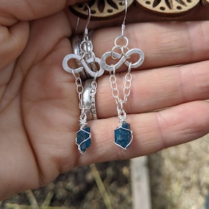 May include: Silver infinity earrings with a dangling blue gemstone wrapped in silver wire.