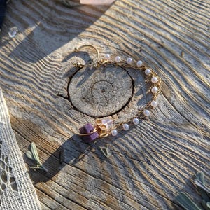 May include: A gold-colored ear cuff with a chain of small white pearls and a cluster of purple and clear crystals. The ear cuff is resting on a weathered wooden surface, with a soft, natural light illuminating the piece.