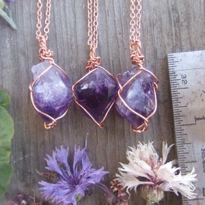 May include: Three amethyst crystal pendants wrapped in copper wire, hanging from delicate copper chains. The pendants are approximately 1 inch in length and are displayed against a wooden background.