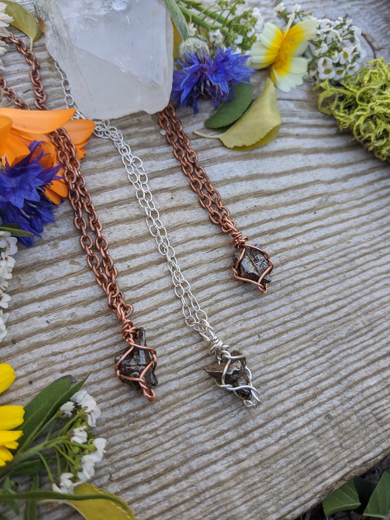 Puede incluir: Tres collares con piedras negras envueltas en alambre. Los collares est&aacute;n sobre una superficie de madera con flores y vegetaci&oacute;n. Los collares tienen cadenas de cobre, plata y oro.
